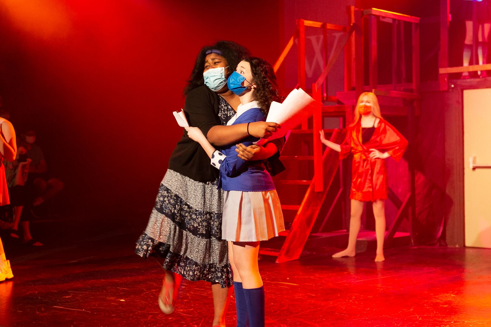 Two teen actors wearing masks hug on stage