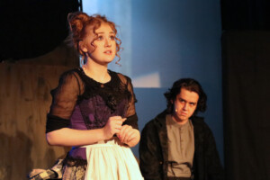 Teenage girl in a black dress and apron looks into the distance while a teenage boy sitting behind her looks at her.