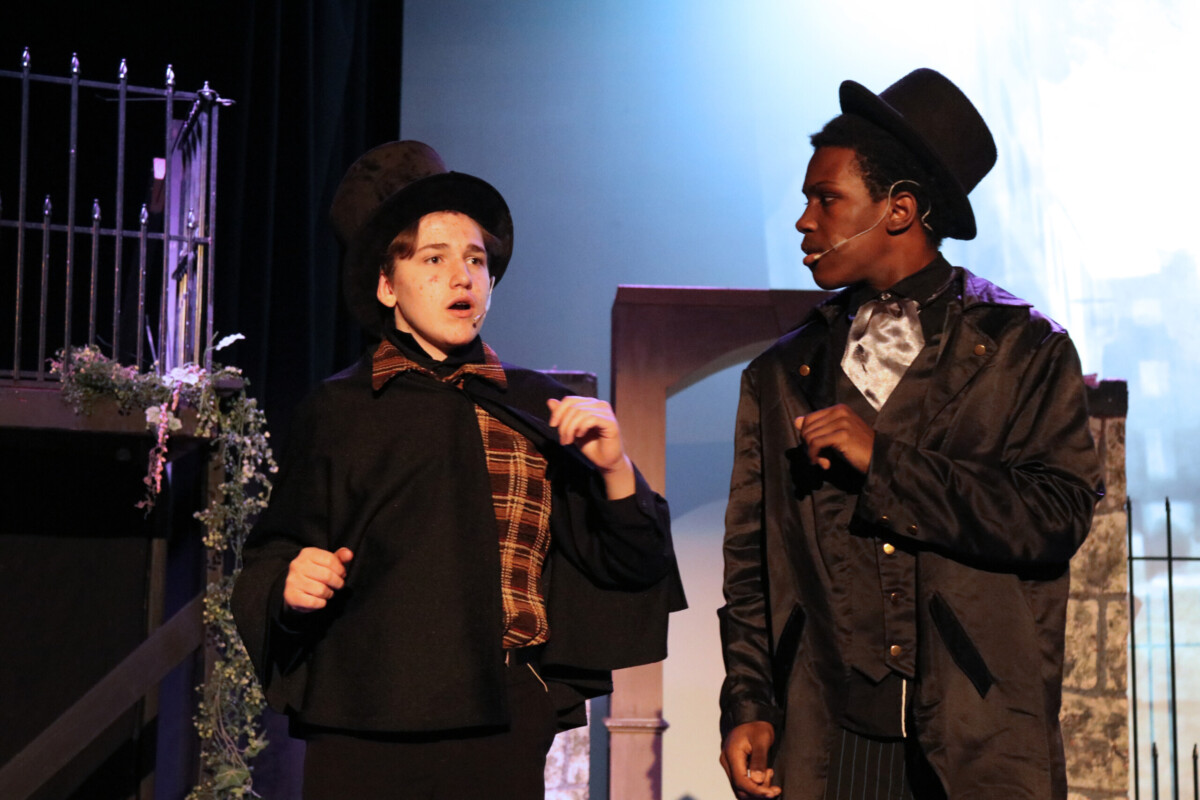 Two academy students perform on stage. Student on the left in a top hat and scarf is speaking while student on the right in a coat and hat looks towards them